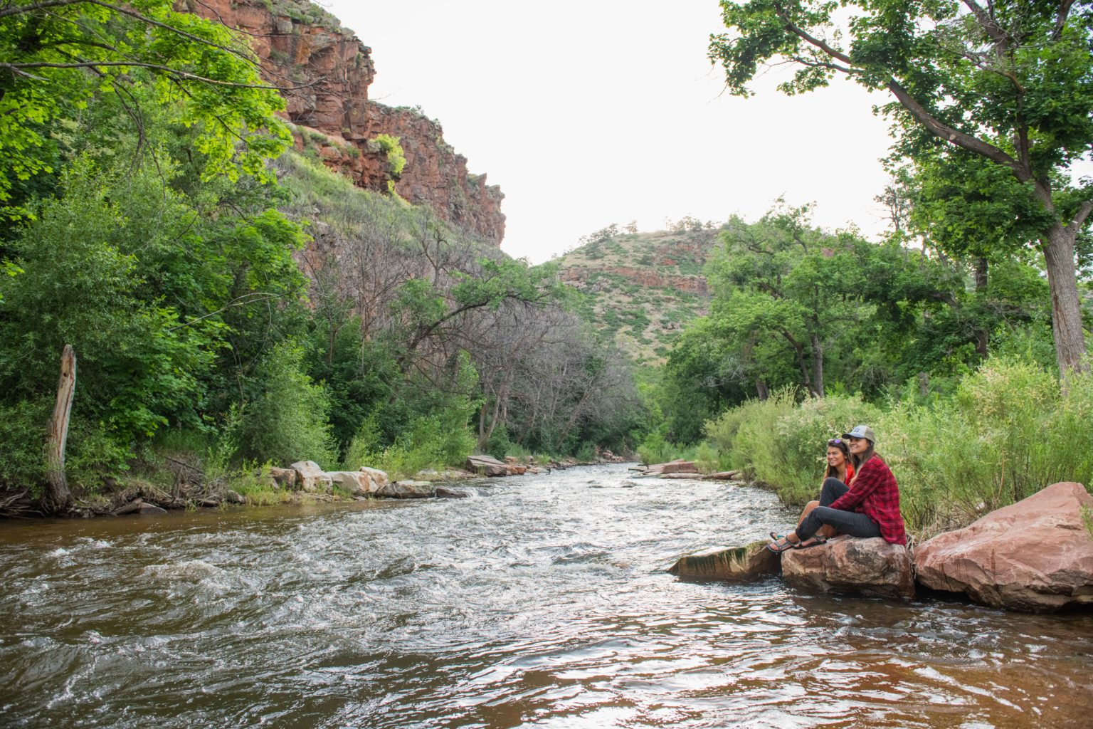 Two women sit on a rock near a stream.