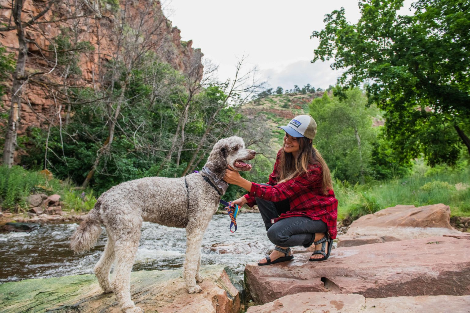 A woman crouches down to pet her dog next to a flowing stream.