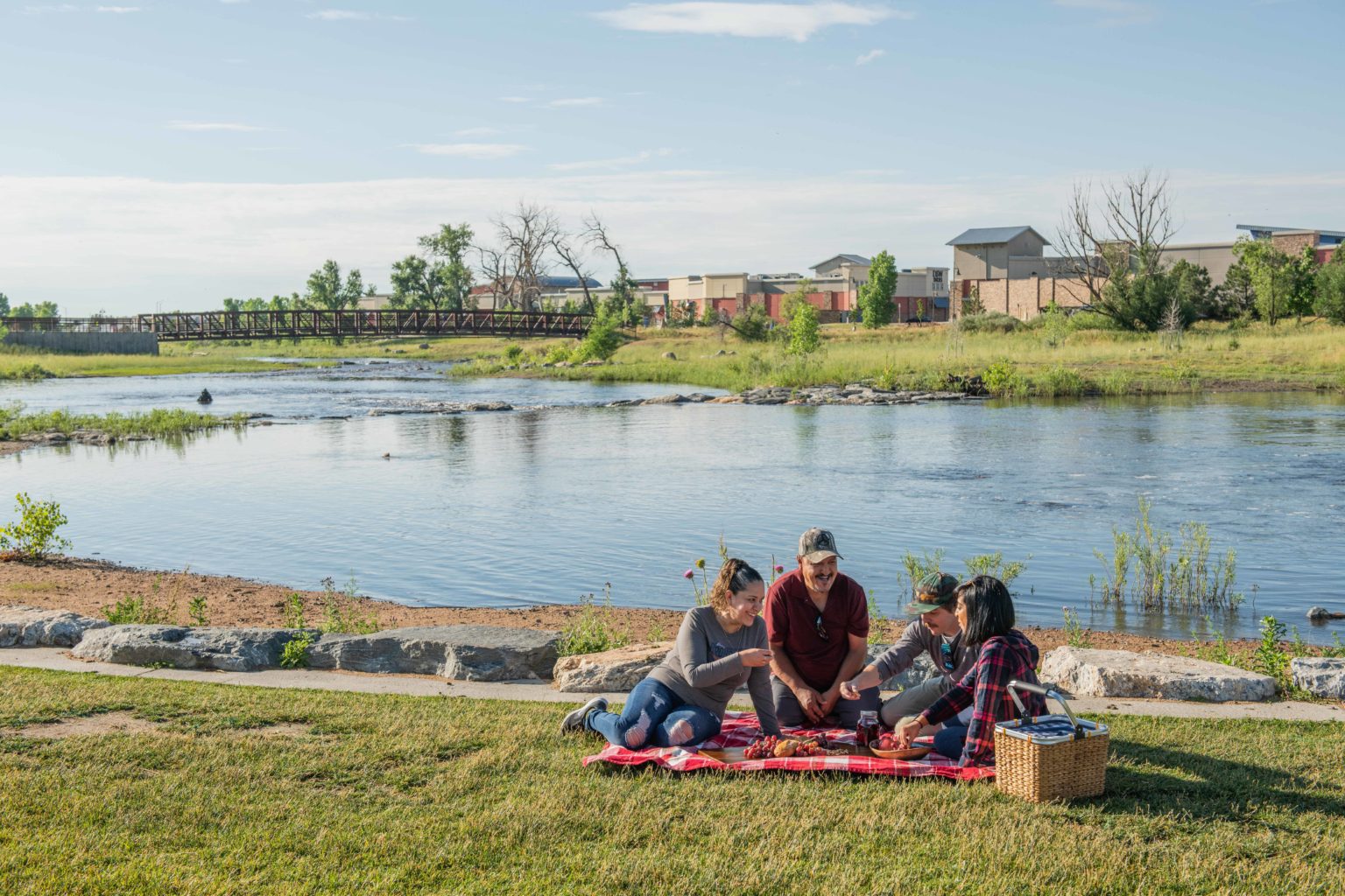 A family of four sit on a red plaid blanket with a picnic basket with a creek and bridge behind them.