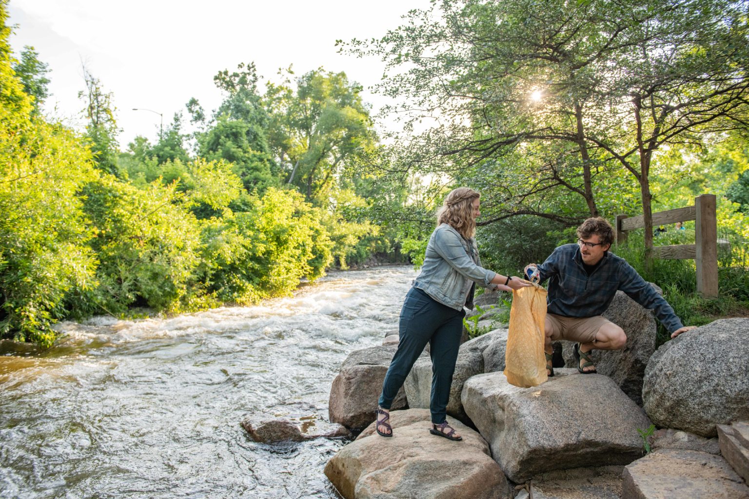 A crouching man places a piece of trash into a trash bag his companion is holding open as they gather litter alongside a fast moving stream.