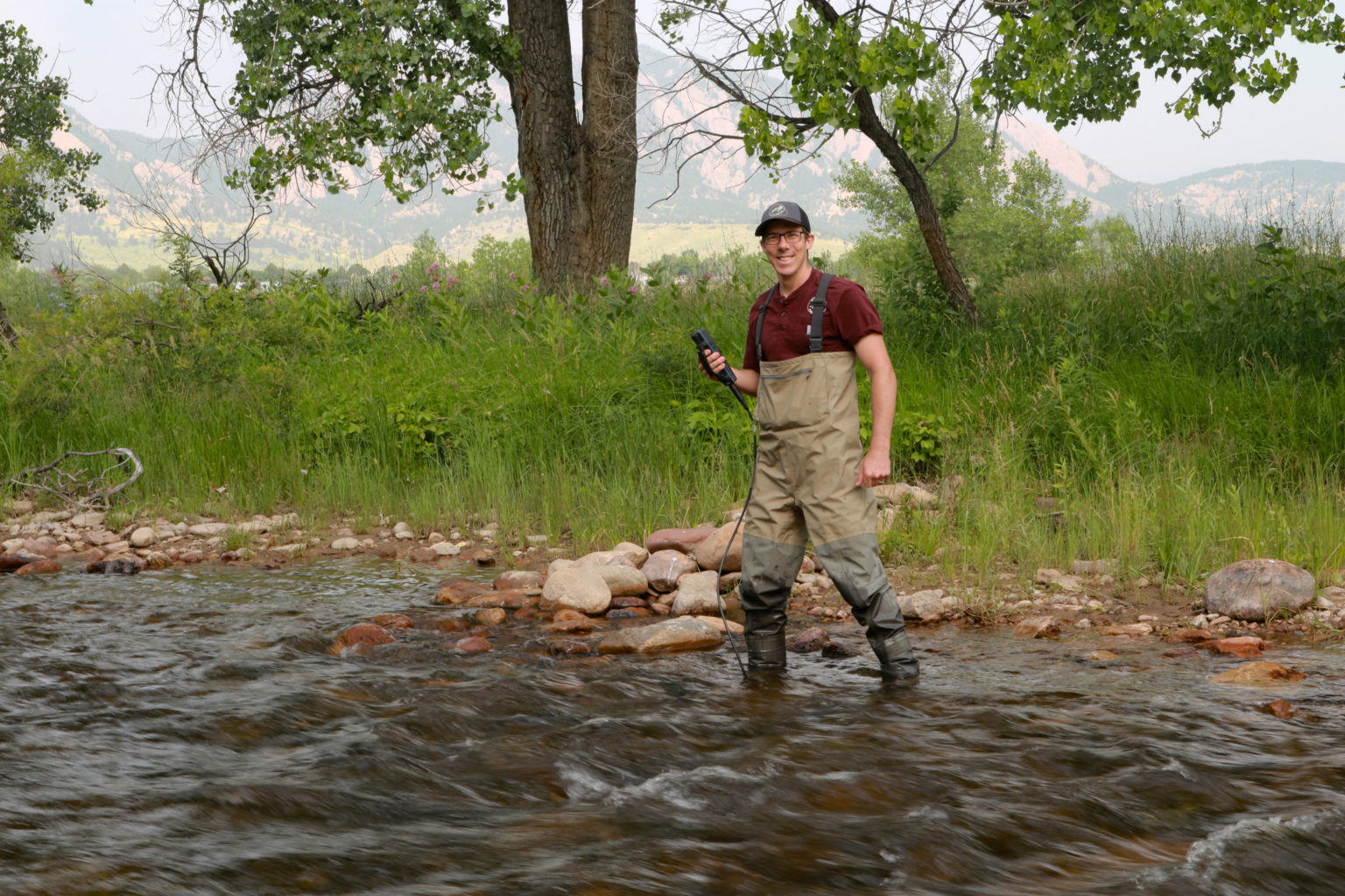 A smiling man holding a water quality testing device stands in a shallow creek.