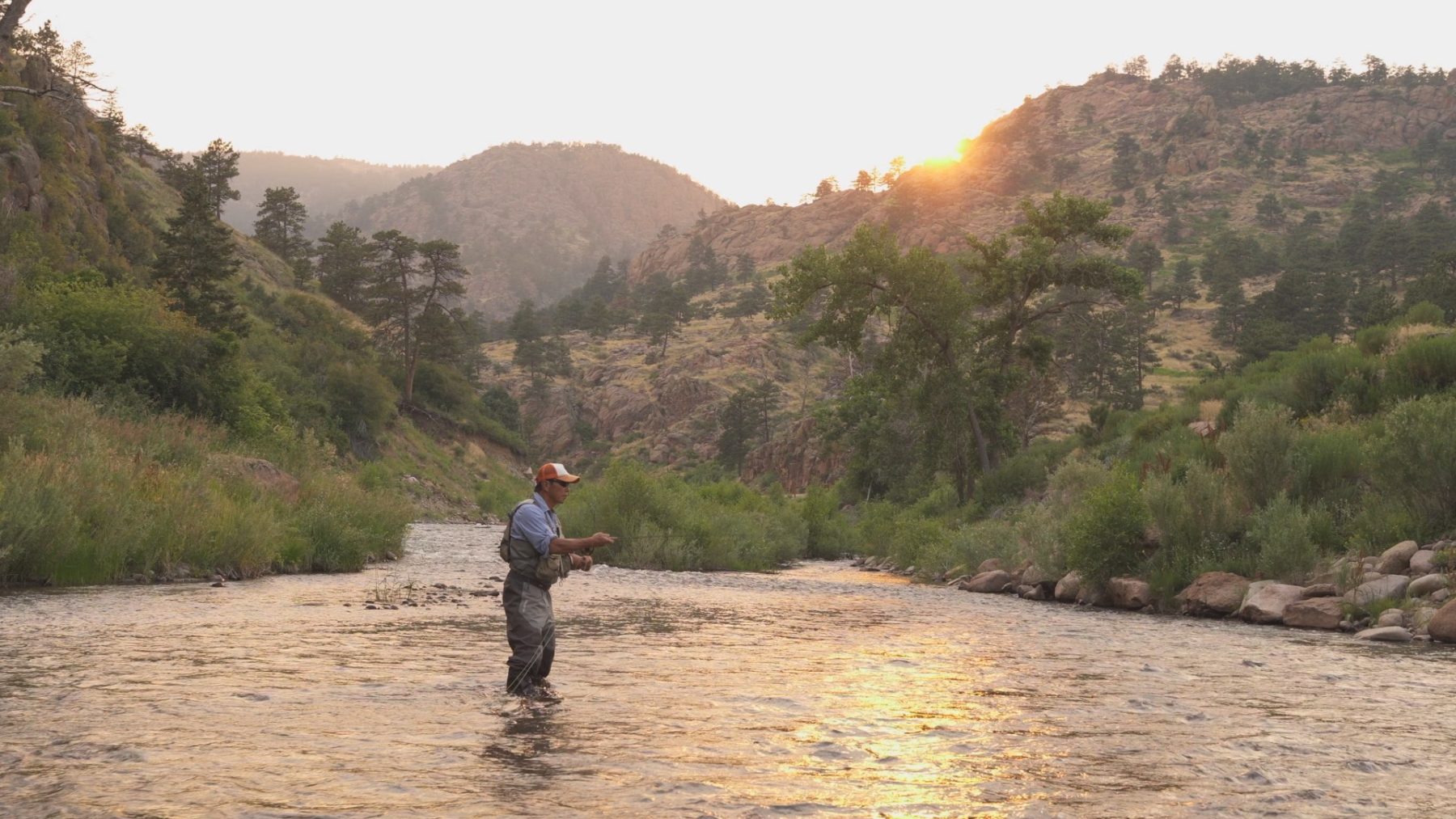 A man stands in the middle of a creek fly fishing as the sun sets behind mountains in the background.