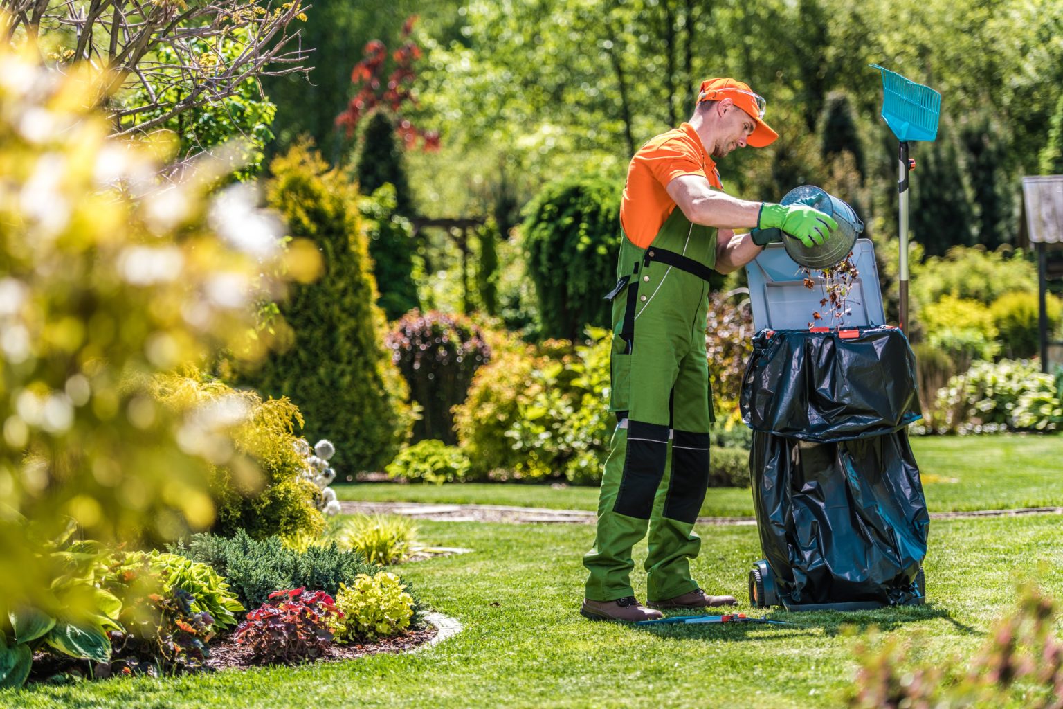 A maintenance person in uniform dumps a bucket of lawn waste into a large trash bag.