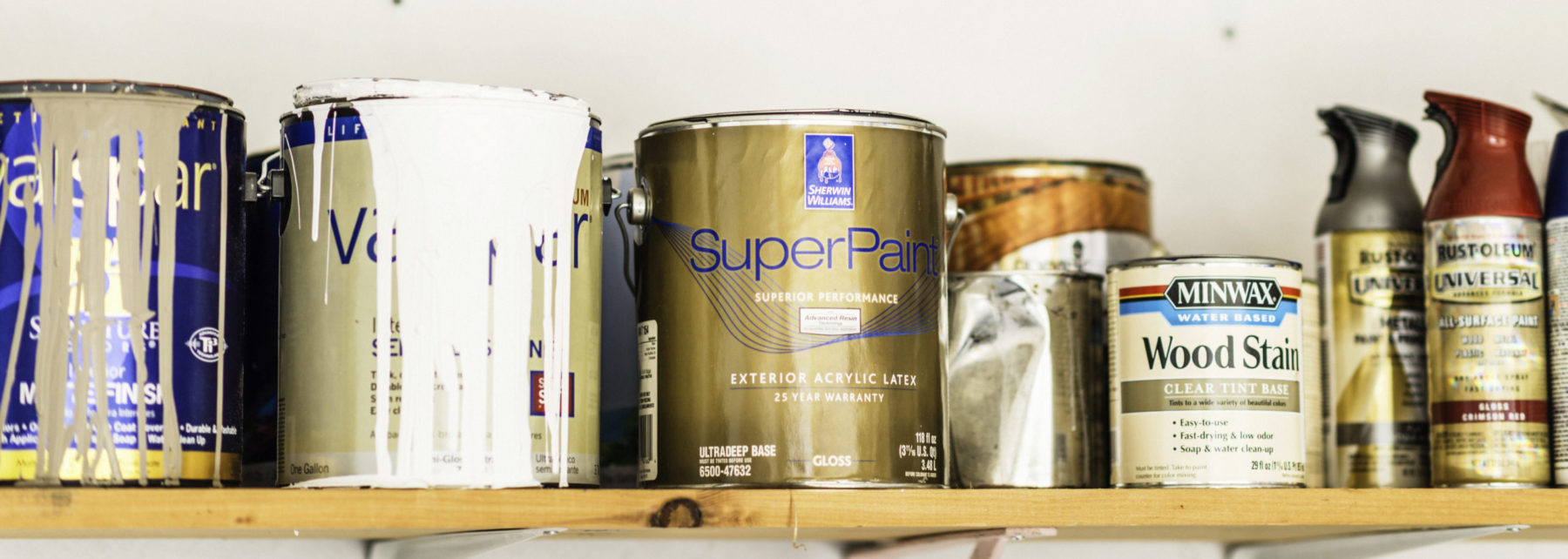 A horizontal shot of a collection of opened and unopened American brand cans of paint, wood stain and paint sprays organized neatly on a wooden shelf.