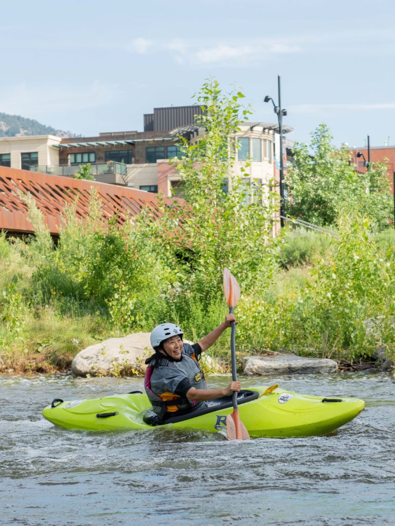 A smiling woman wearing a life vest and helmet paddles down a creek in a kayak.