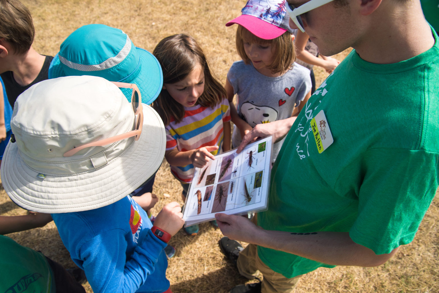 A group of young students gather around a teacher and look at a handout with images of aquatic insects..