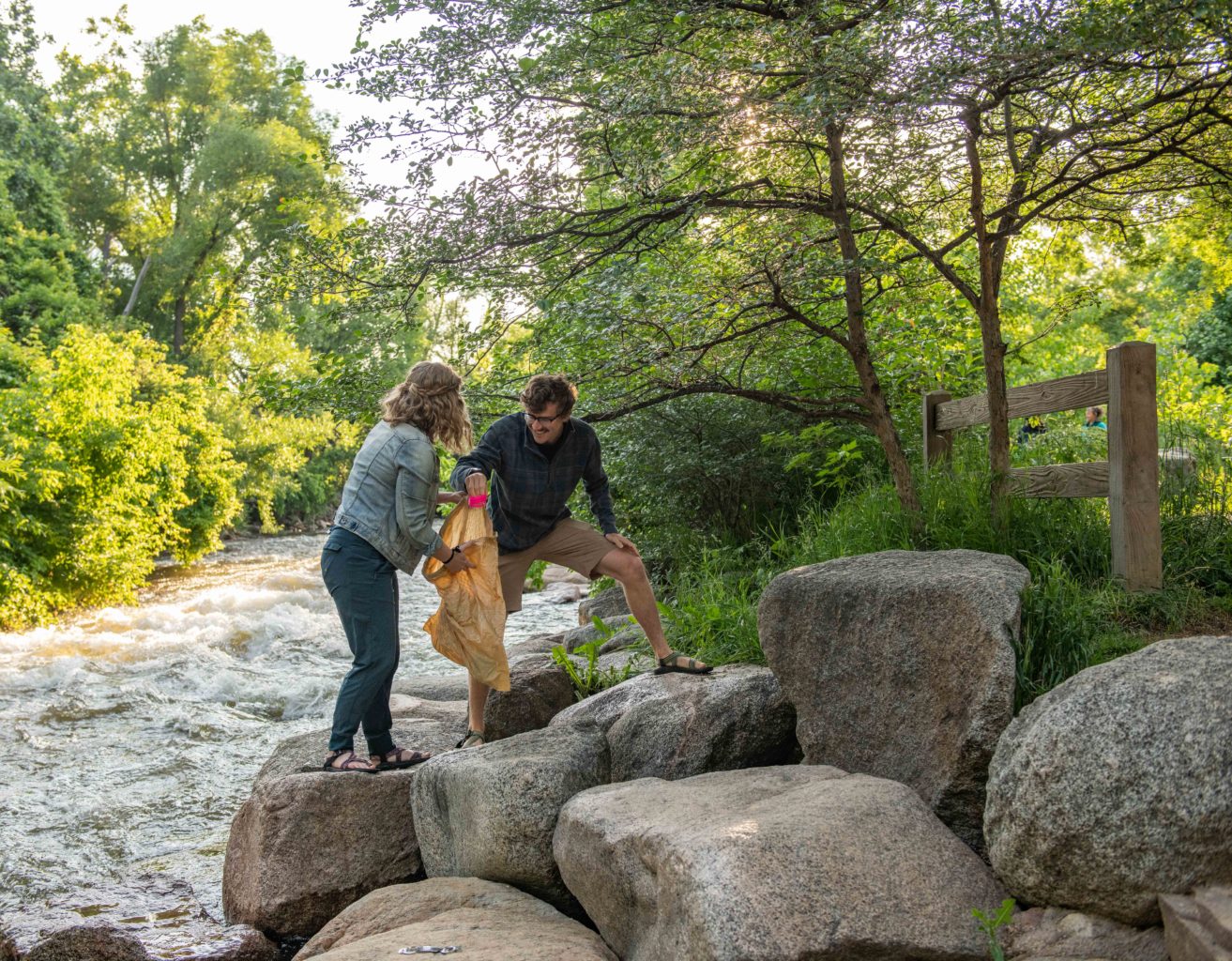 A woman holds open a trash bag as a man places a piece of trash into it. They are cleaning up trash on boulders along a creek.