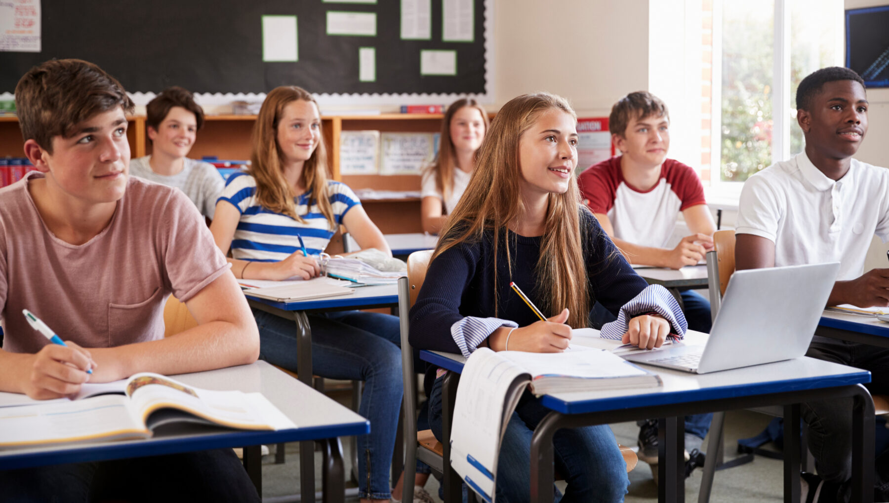 Students sitting at desks in a classroom listening to someone speaking at the front of the classroom.