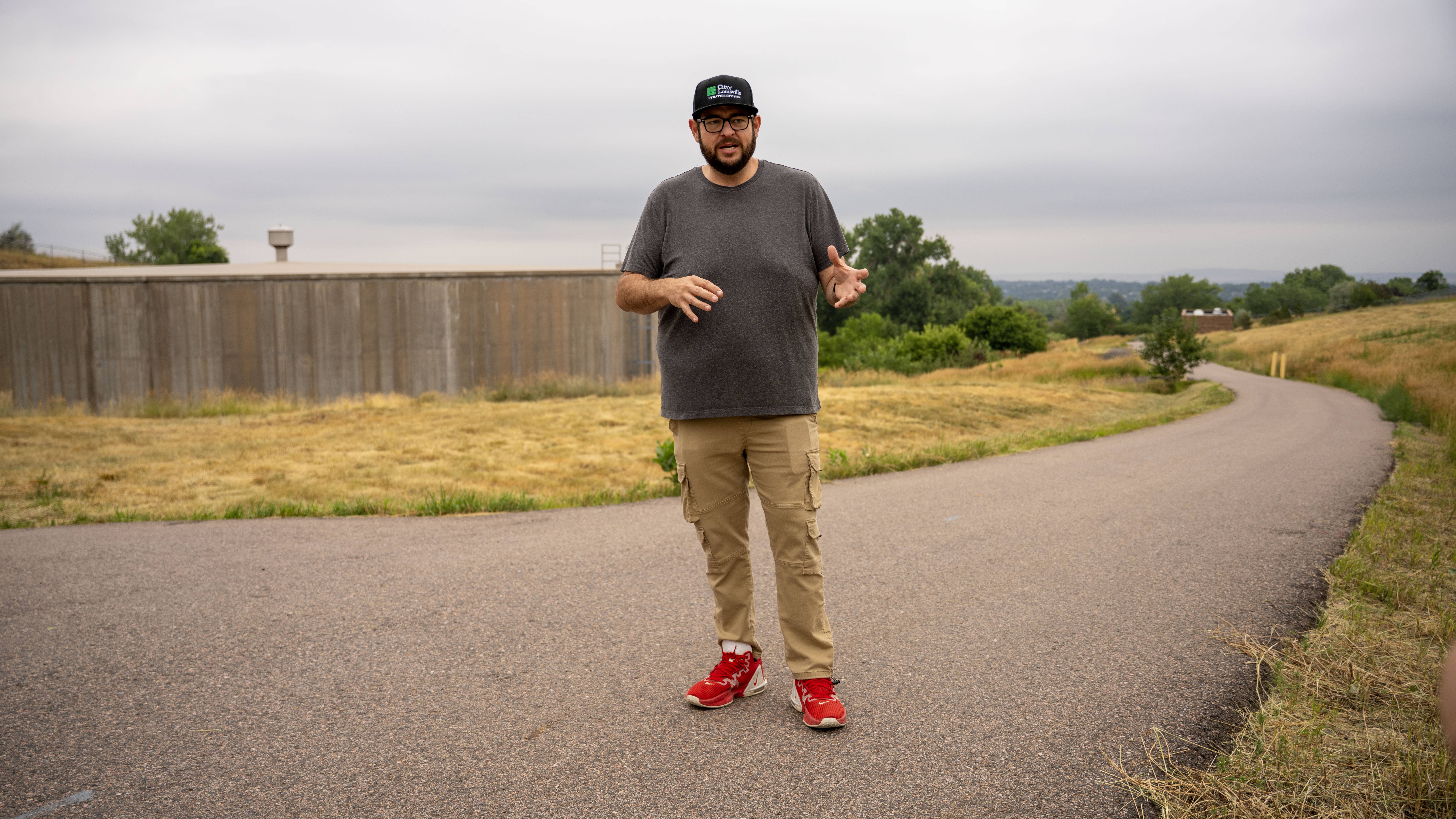 A man stands on a road, talking and motioning with his hands. A large water storage tank is in the background.