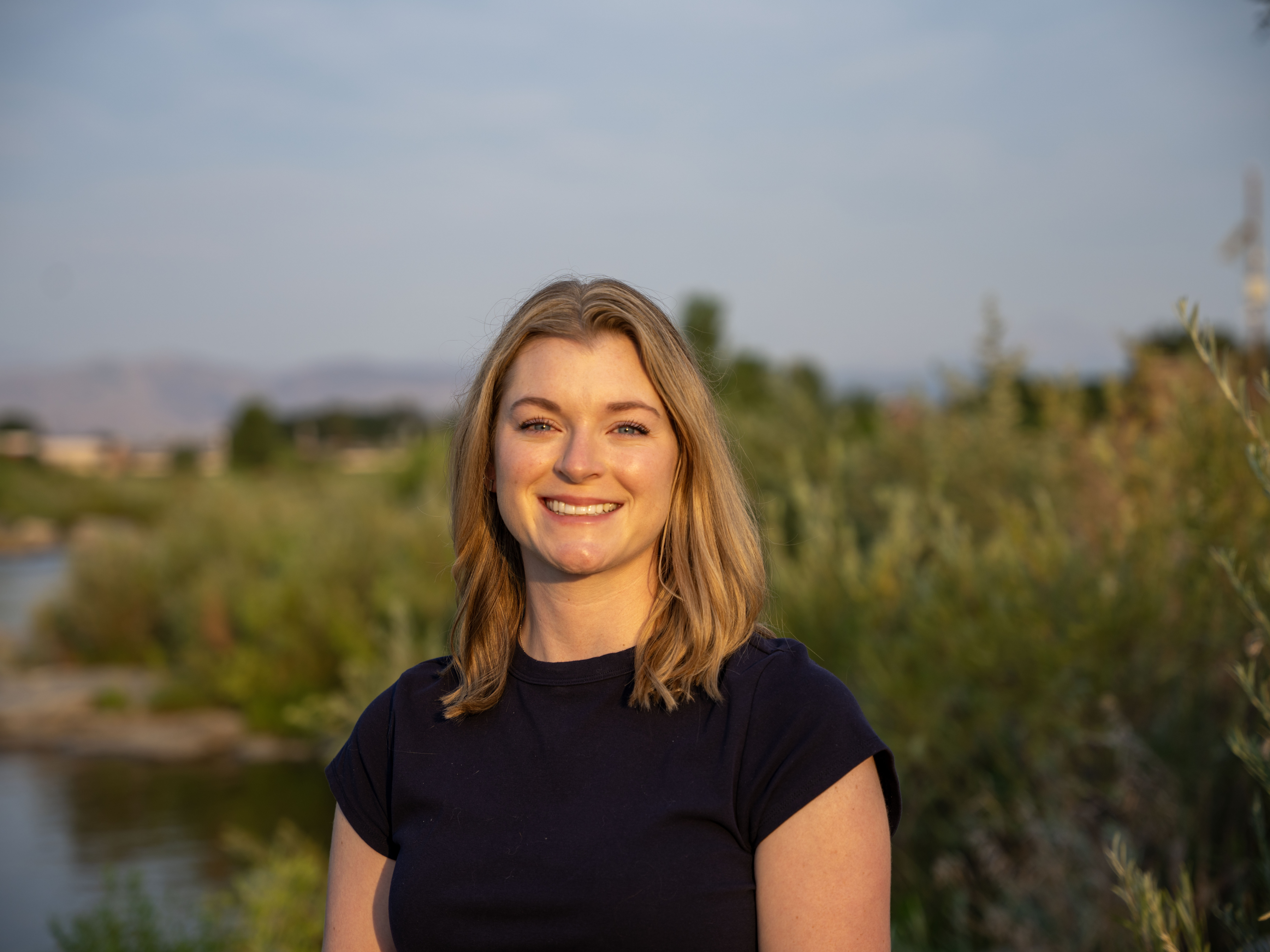 A smiling woman stands on a river bank with vegetation in the background.