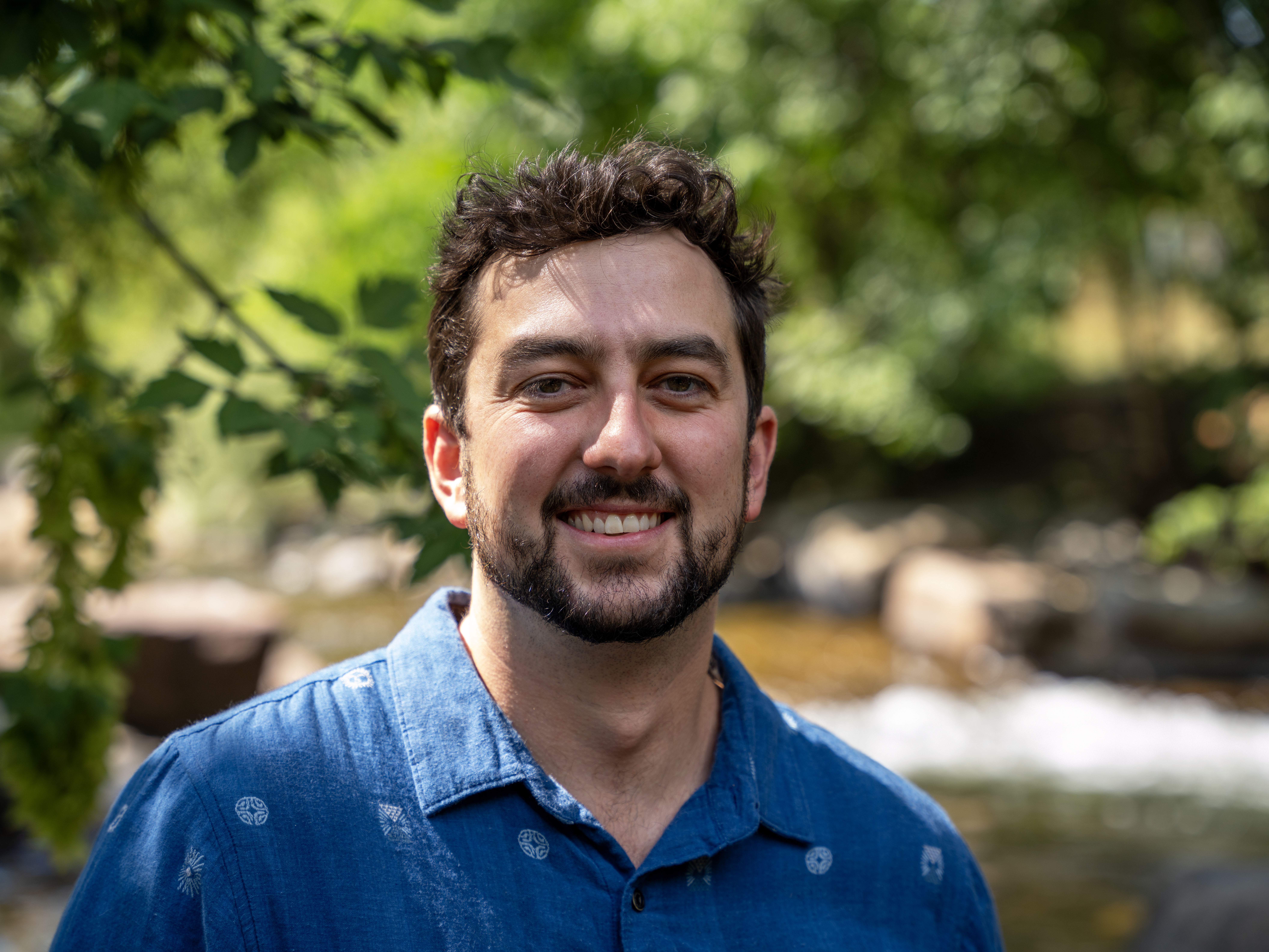 Close up image of a smiling man standing in front of a shaded river.