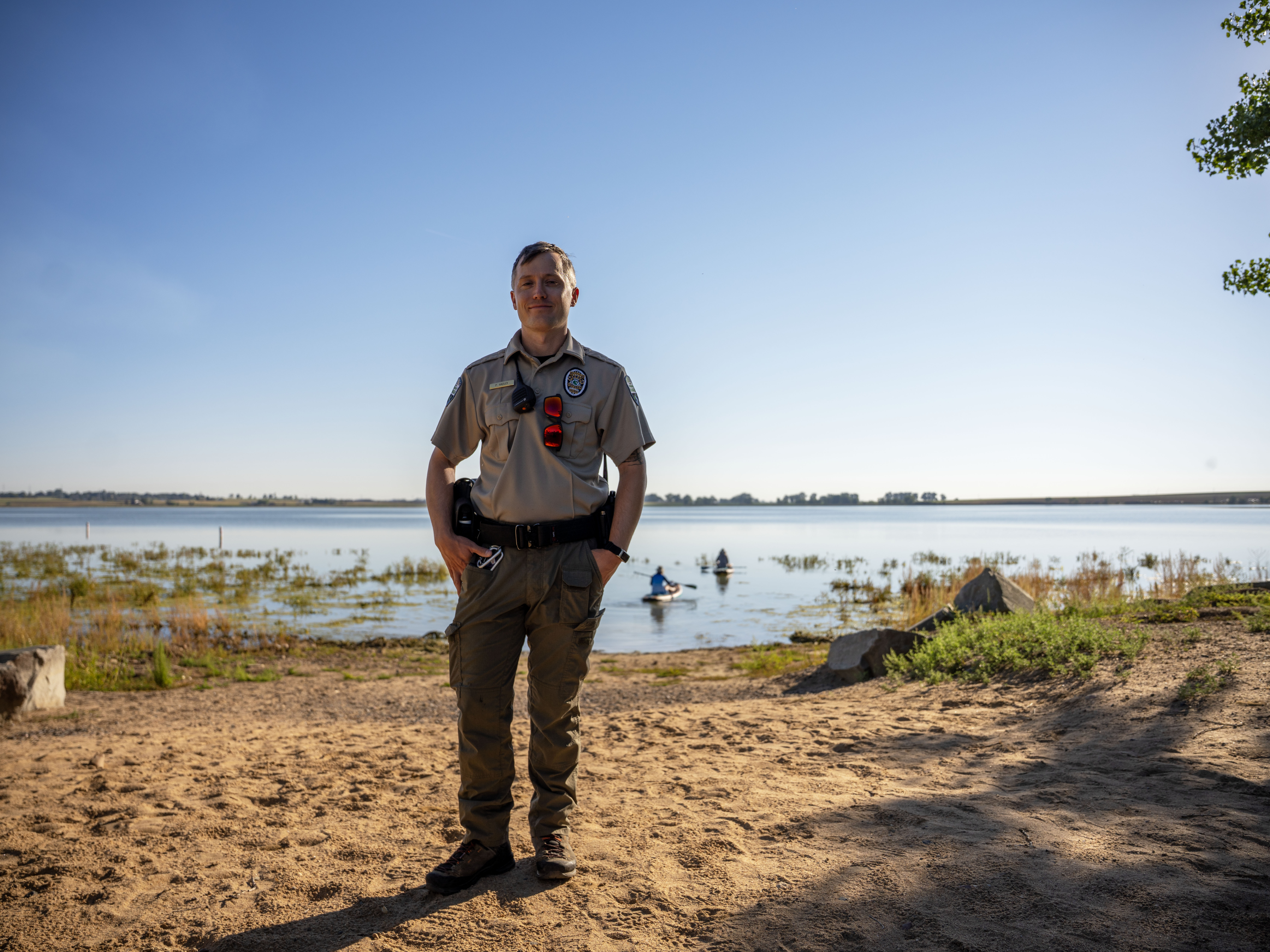 A man in law enforcement uniform stands on the beach of a reservoir.