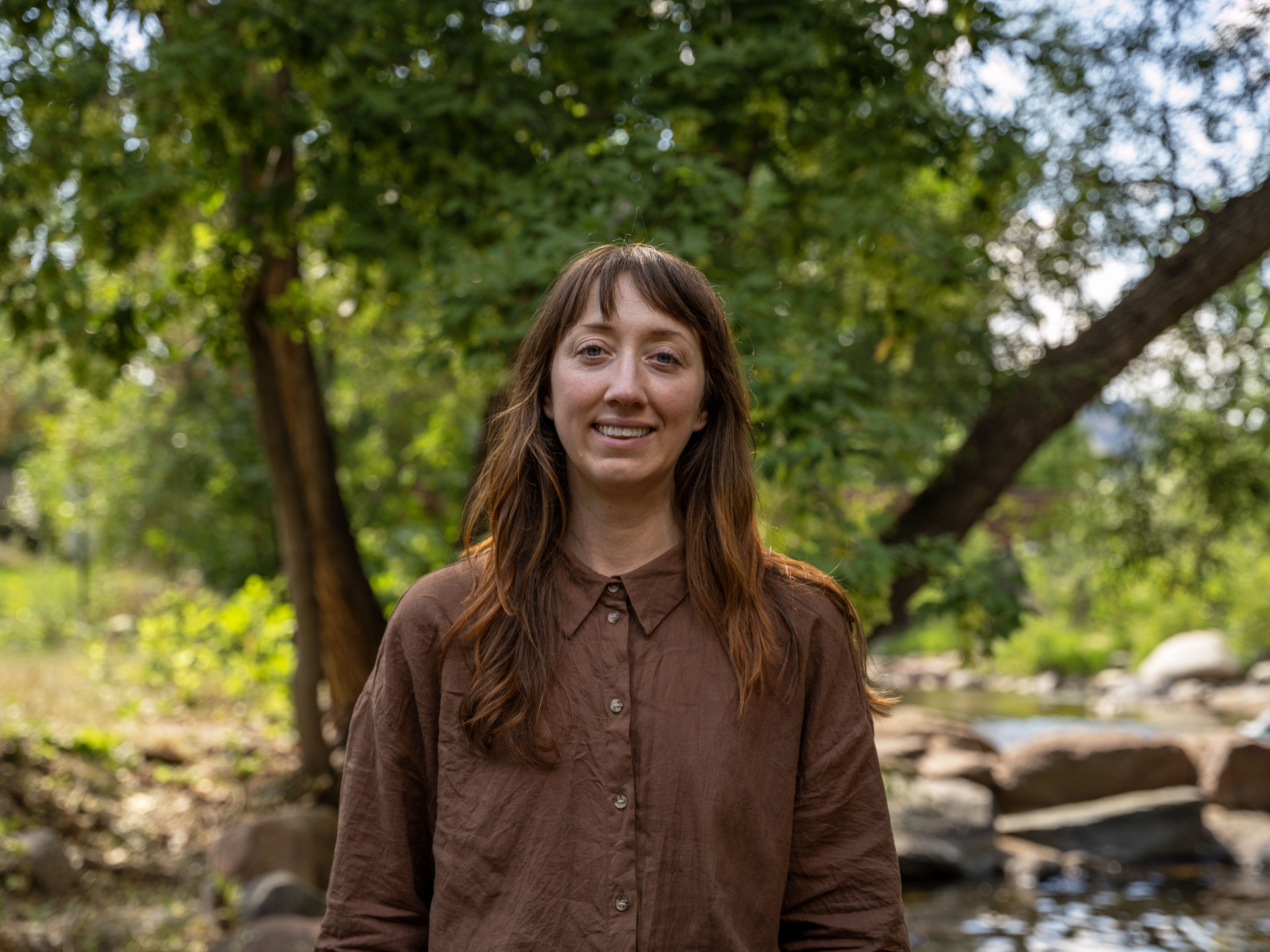 A woman stands outside by a tree and river.