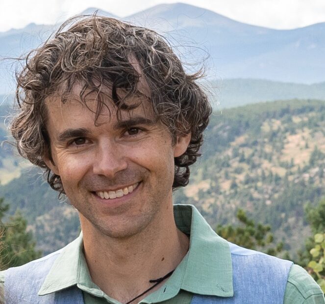 A smiling man standing outdoors with mountains in the background.